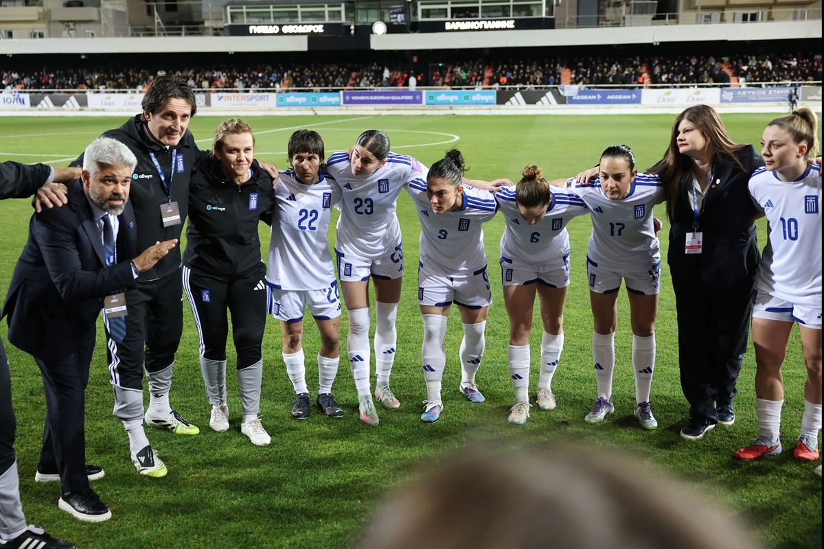 Ein weibliches Fußballteam in weißen Trikots versammelt sich zum Team-Besprechung auf dem Feld, Trainer steht daneben.