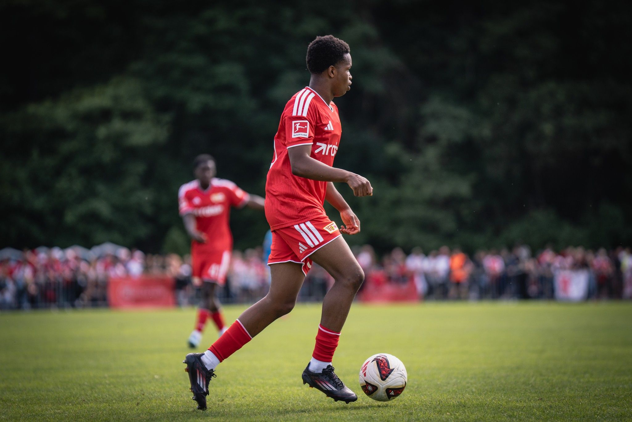 Ein junger Fußballspieler in rotem Trikot dribbelt den Ball auf einem Rasenfeld, Zuschauer im Hintergrund.