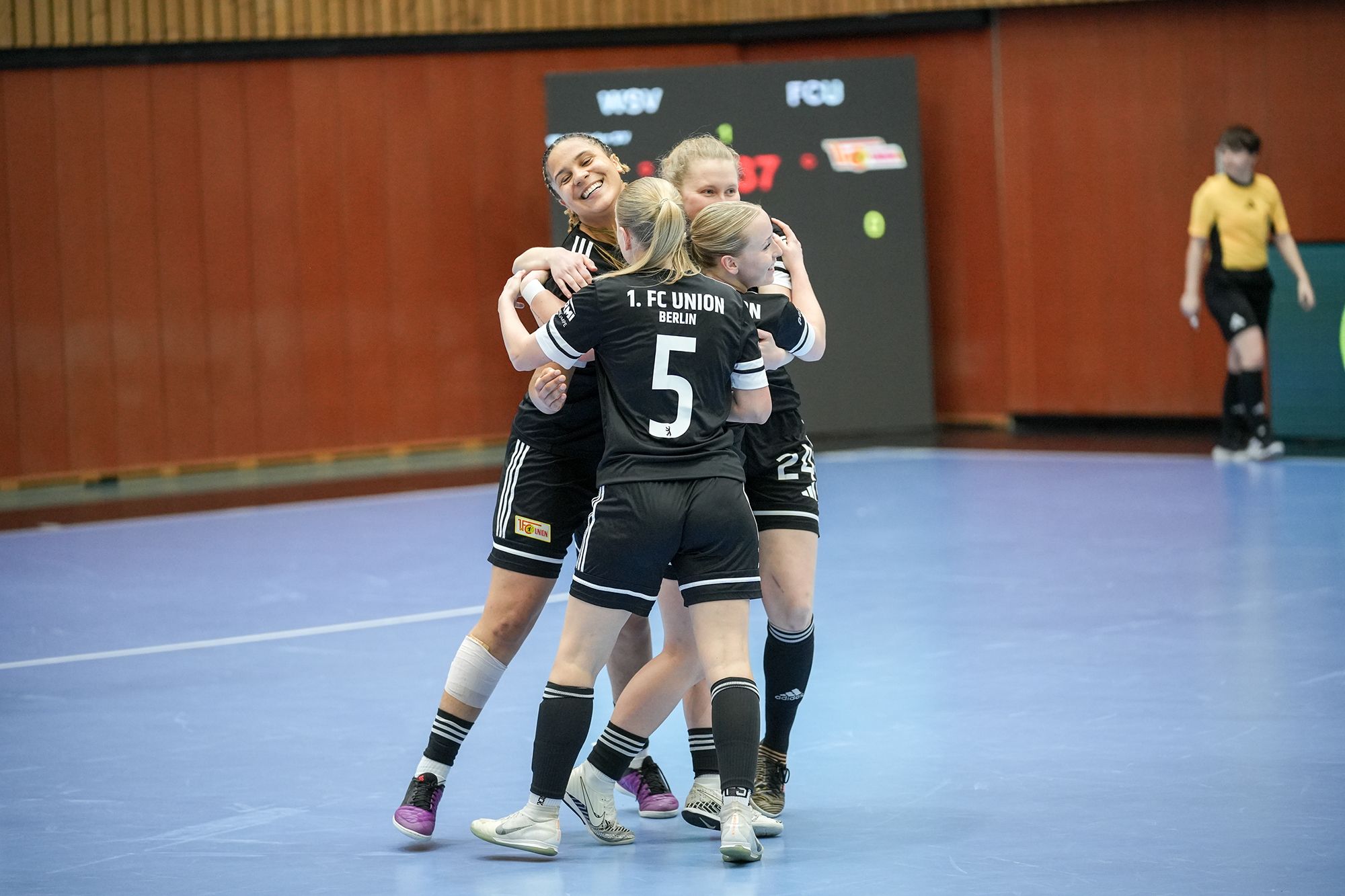 Drei weibliche Spielerinnen in schwarzen Sporttrikots umarmen sich jubelnd in einer Sporthalle nach einem Erfolg.