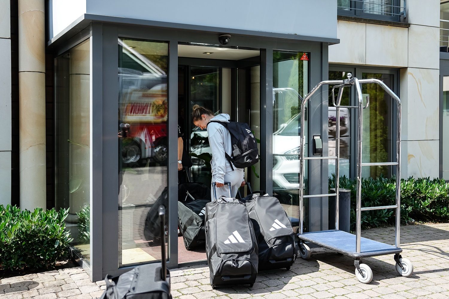 Eine Person mit einem Rucksack beugt sich über Gepäckstücke vor einem Hotel mit einem Gepäckwagen im Hintergrund.