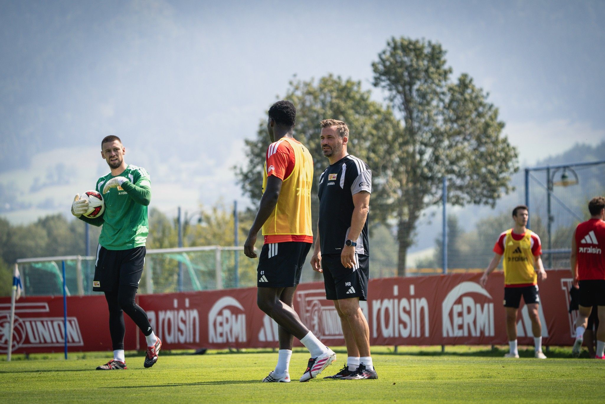 Training der Fußballmannschaft im Freien, mit Spielern und Trainern auf dem Platz vor einer Berglandschaft.
