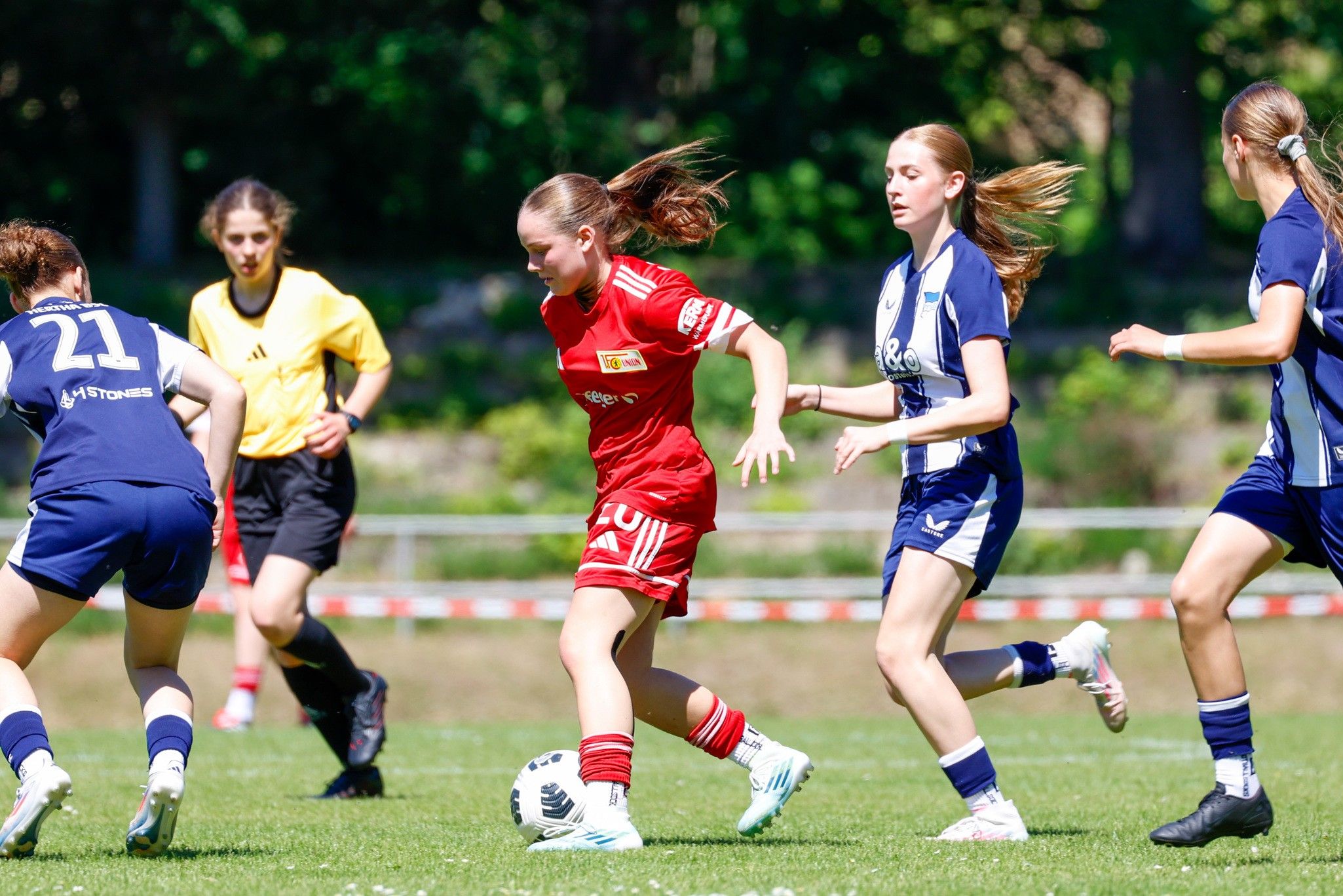 Spielerinnen in roten und blauen Trikots auf einem Fußballfeld während eines Spiels. Eine Spielerin dribbelt den Ball.