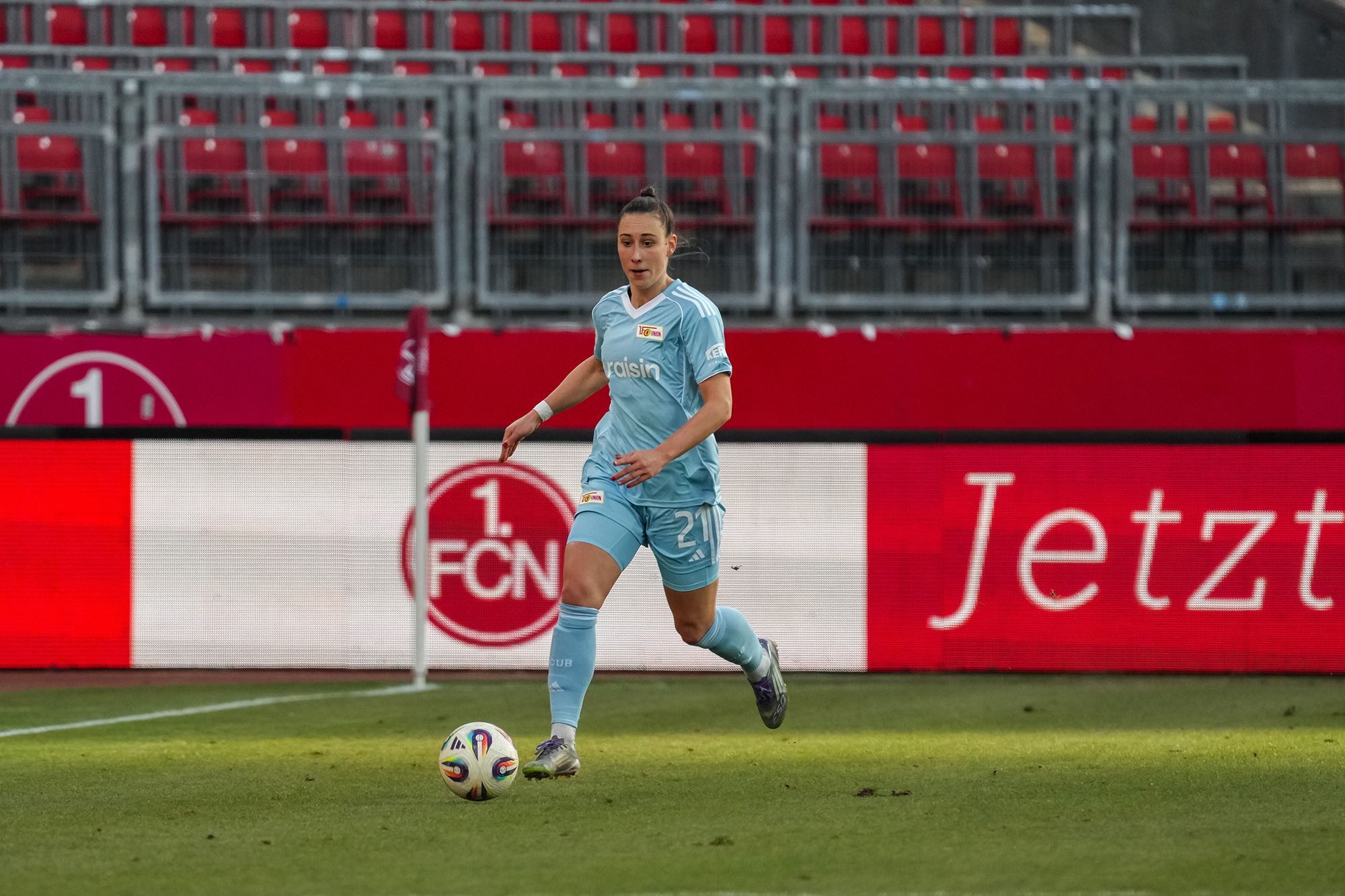 Eine Fußballspielerin in hellblauem Trikot läuft auf dem Feld, während das Stadion im Hintergrund sichtbar ist.