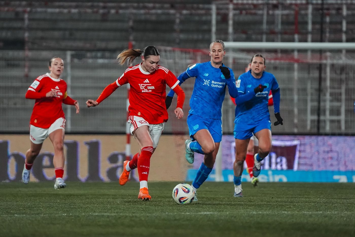Eine Spielerin in rotem Trikot dribbelt den Ball, während zwei Spielerinnen in blauen Trikots sie verfolgen.