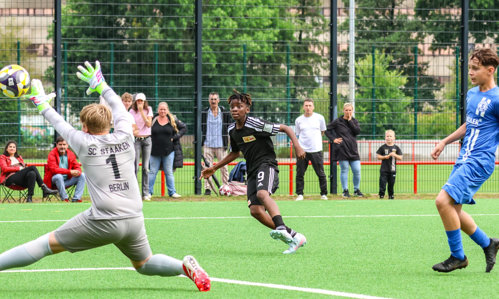 Ein Fußballspiel auf einem Kunstrasenplatz, ein Spieler schießt auf das Tor, während der Torwart reagiert und Zuschauer im Hintergrund stehen.