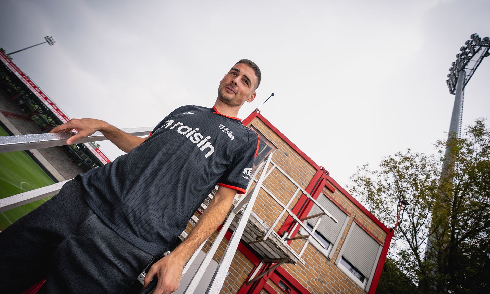 Ein Mann in einem schwarzen Fußballtrikot steht auf einer Treppe vor einem Stadion bei bewölktem Himmel.