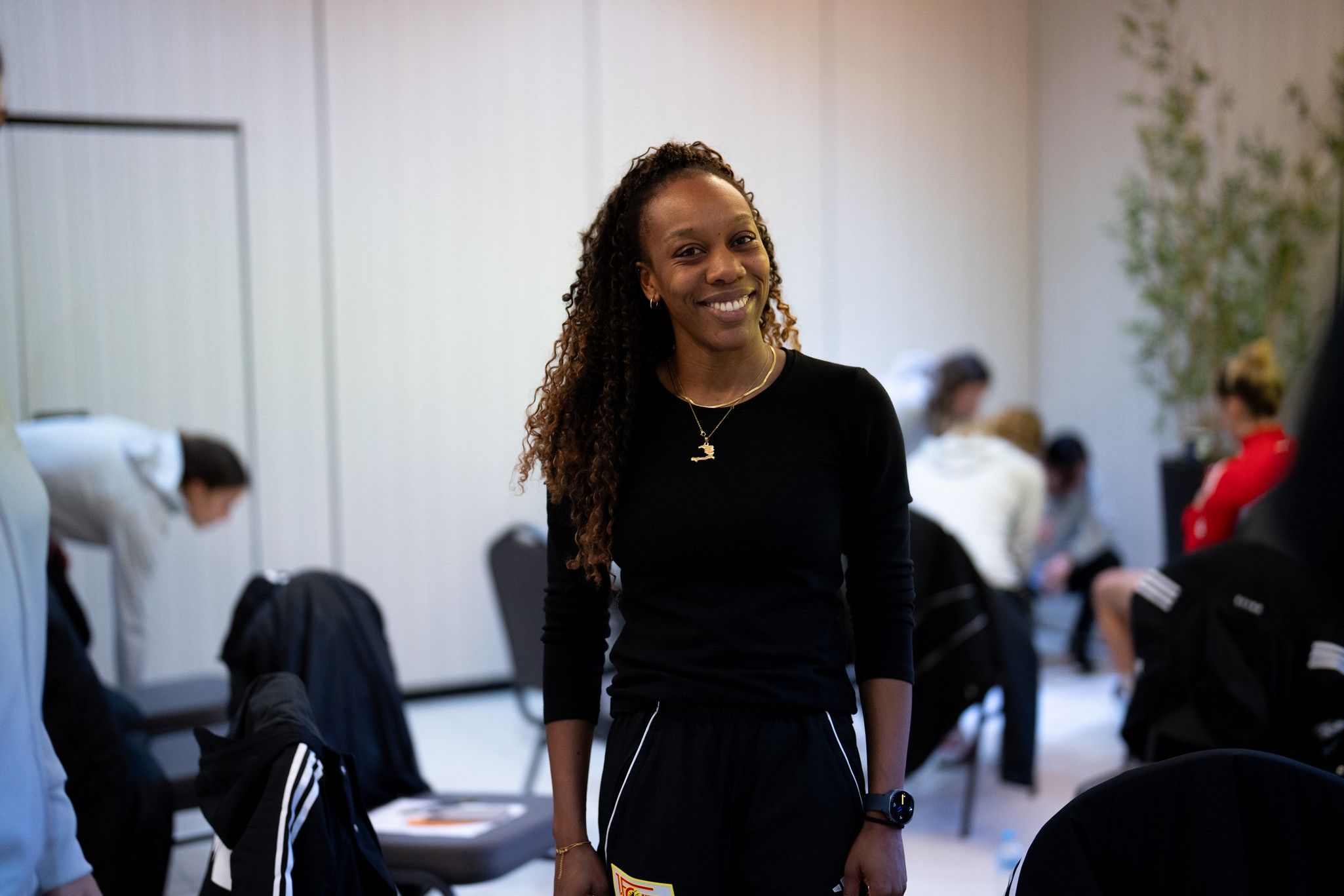Eine lächelnde Frau mit lockigem Haar steht in einem Raum mit Stühlen, während andere Personen im Hintergrund beschäftigt sind.