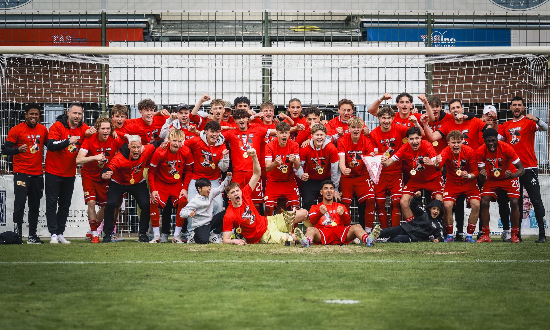 Gruppe von jubelnden jungen Fußballspielern in roten Trikots mit Medaillen, posierend vor einem Tor.