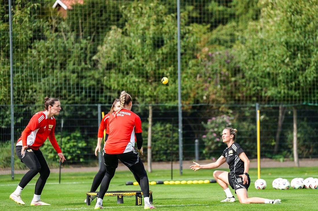 Vier Frauen in Sportkleidung trainieren auf einem Fußballplatz, während eine von ihnen einen Ball wirft.