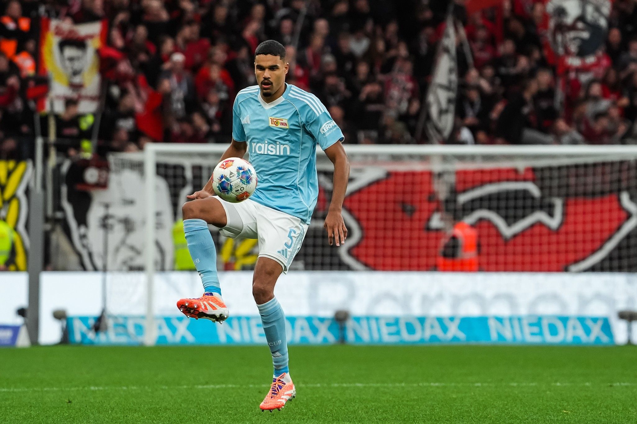 Ein Fußballspieler in hellblauem Trikot kontrolliert den Ball auf dem Platz vor einer jubelnden Menge.