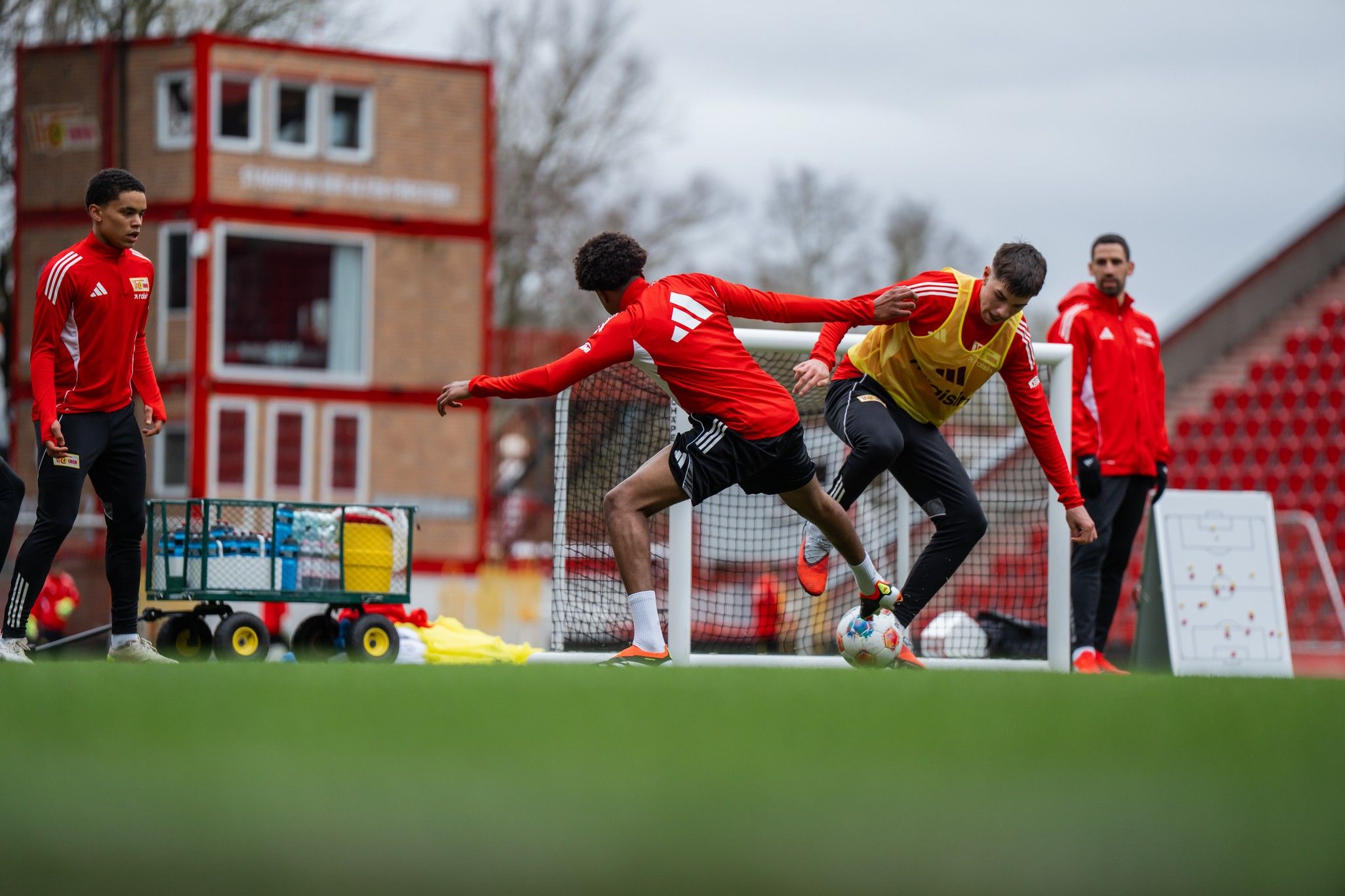 Zwei Fußballspieler in Trainingskleidung üben auf einem Platz, während ein dritter Spieler und ein Trainer zuschauen.
