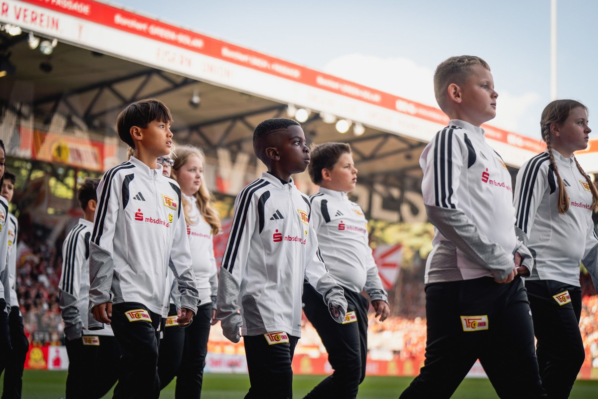 Kinder in weißen Trikots gehen auf einem Fußballfeld, im Hintergrund ein Stadion voller Fans.