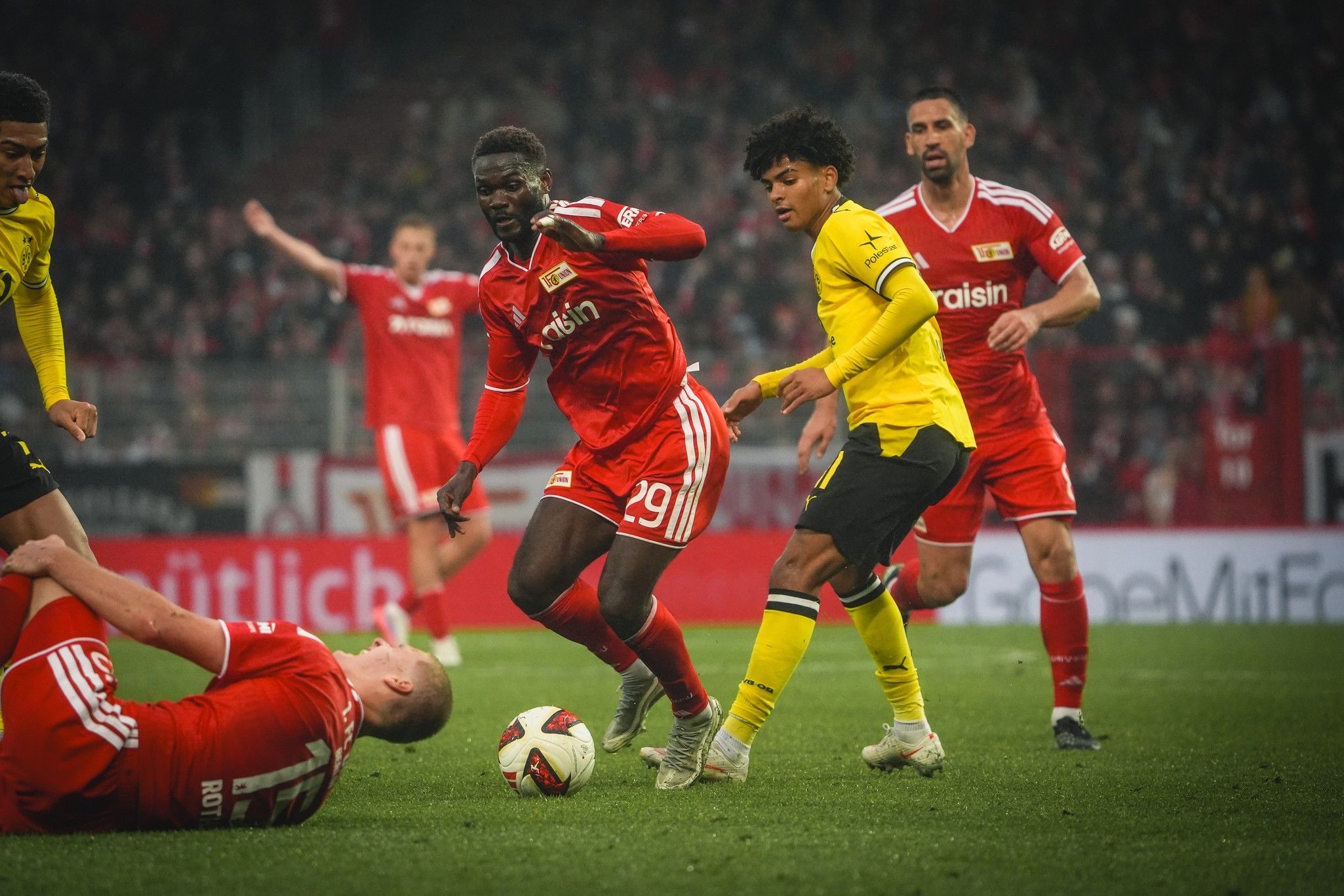 Spielszene aus einem Fußballspiel mit Spielern in roten und gelben Trikots, ein Spieler fällt hin, während andere aktiv sind.