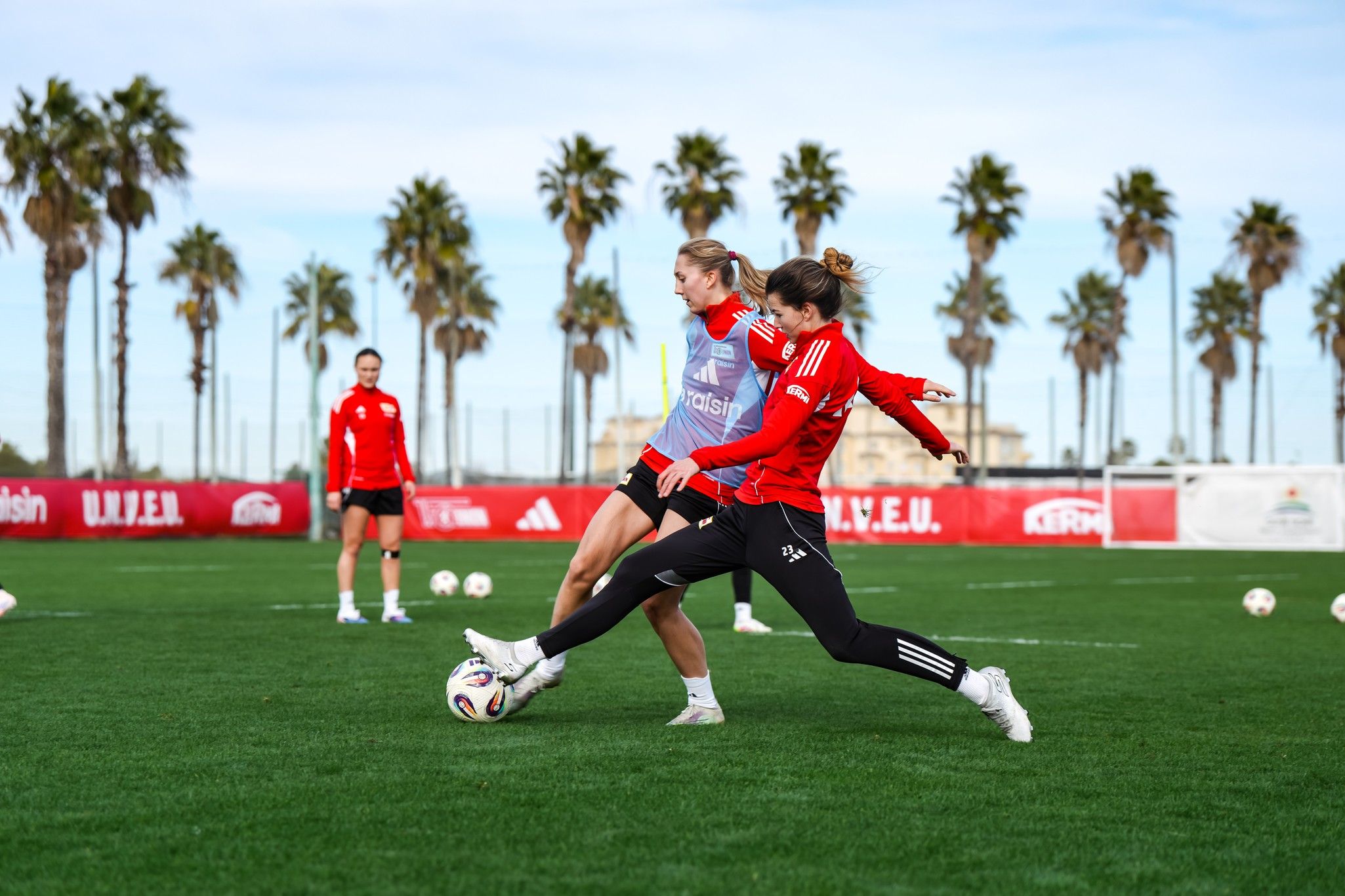 Zwei Fußballspielerinnen in roten Trainingsanzügen treten während eines Trainings auf einem grünen Platz gegen den Ball. Palmen im Hintergrund.