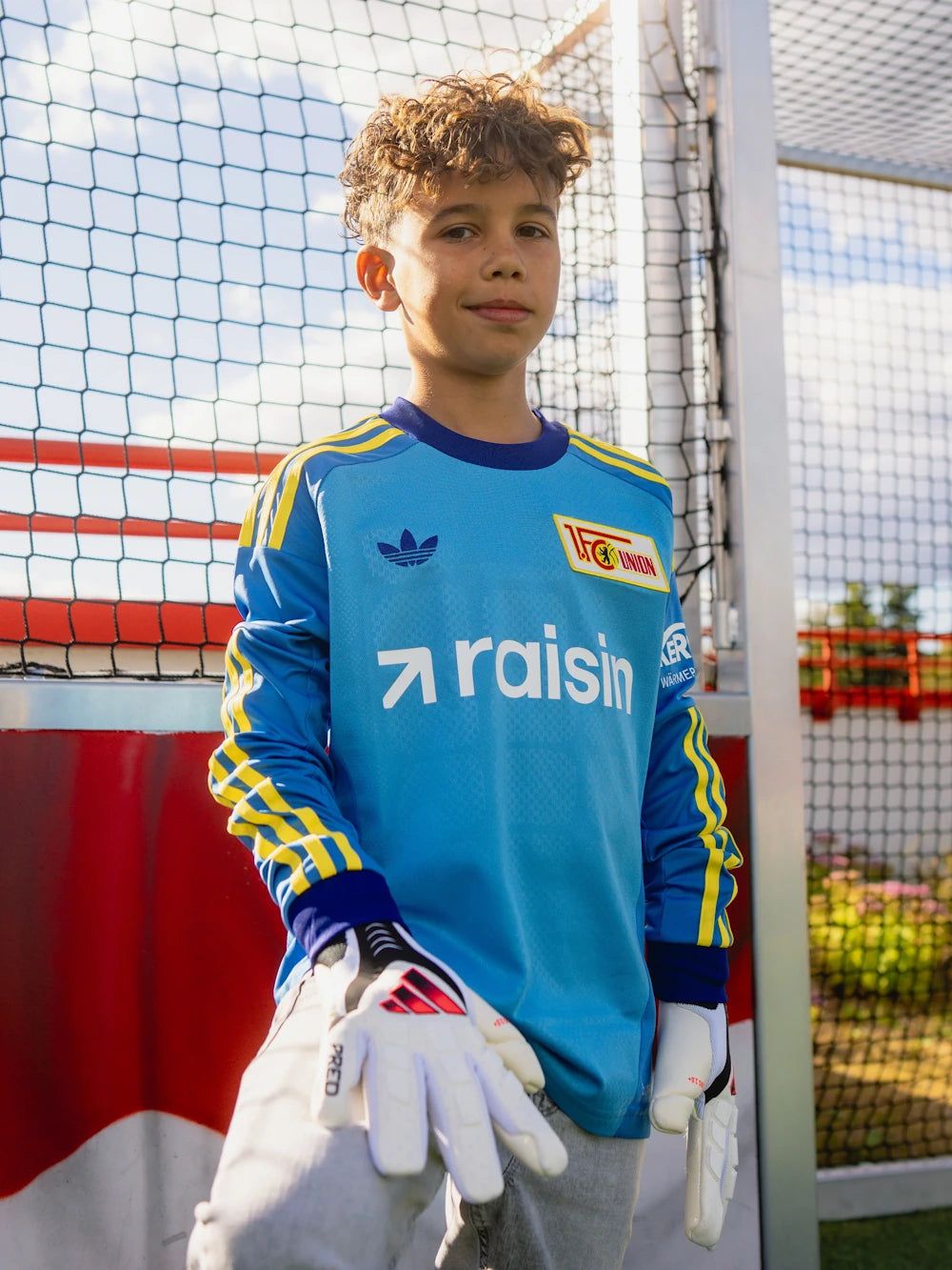 Ein junger Torwart in einem blauen Trikot mit der Aufschrift "raisin" und Schutzhandschuhen steht vor einem Fußballtor.