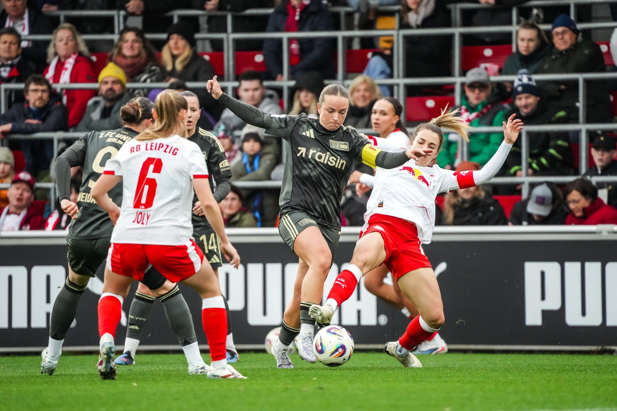 Spielszene im Frauenfußball: Spielerinnen kämpfen um den Ball in einem intensiven Wettkampf vor jubelndem Publikum.