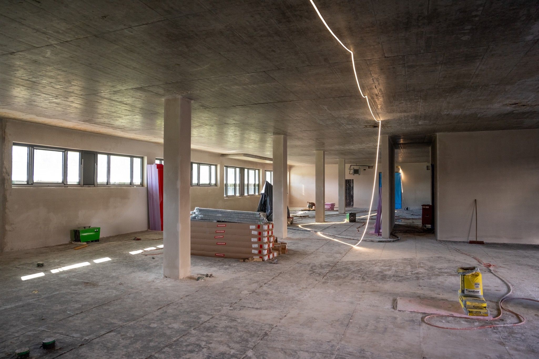 Blick in einen unvollendeten Raum mit Betonboden, offenen Wänden und Baustellenmaterialien. Tageslicht fällt durch die Fenster.