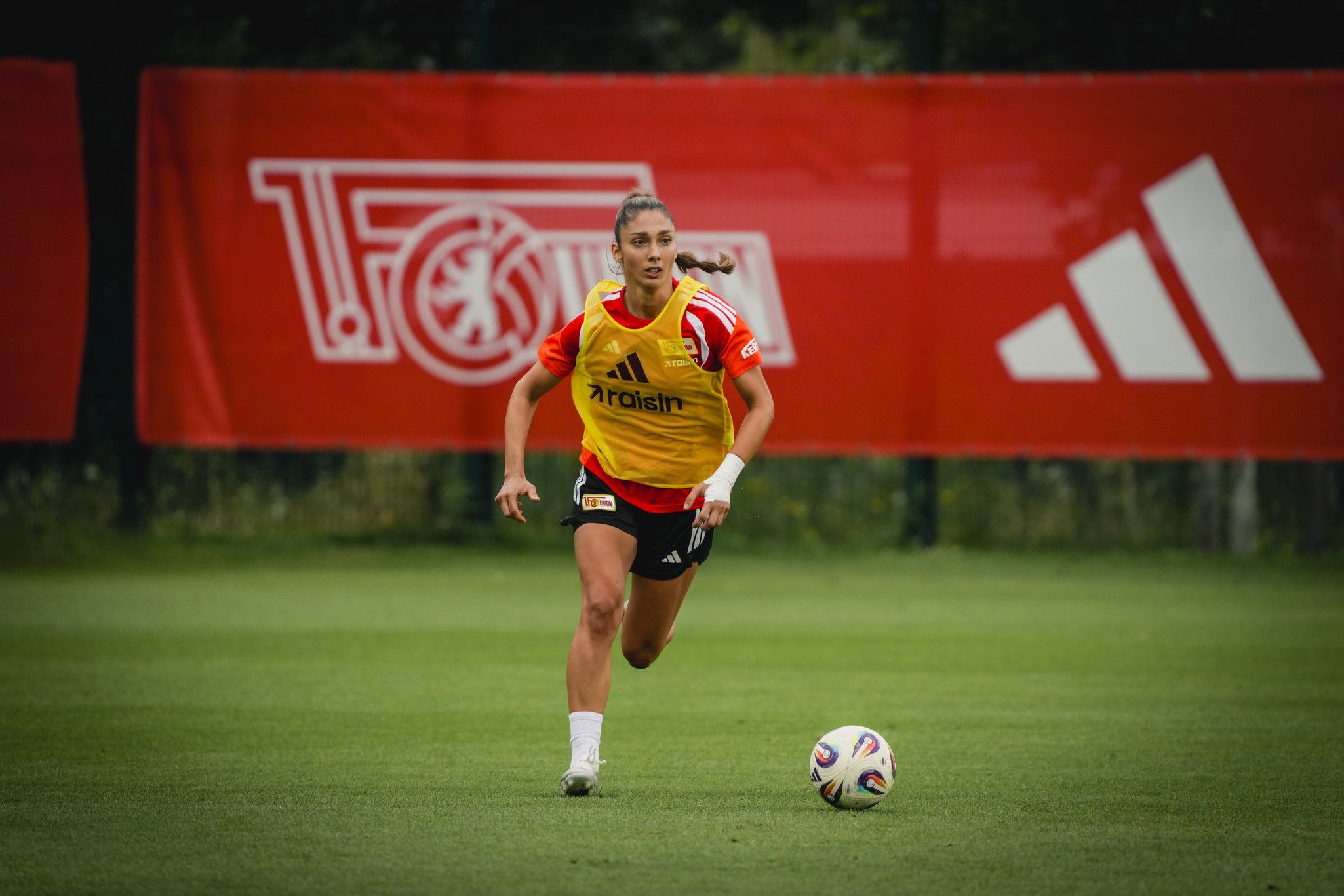 Eine Sportlerin in gelber Trainingskleidung sprintet auf einem grünen Fußballfeld mit einem Ball vor sich. Im Hintergrund sind Werbebanner zu sehen.