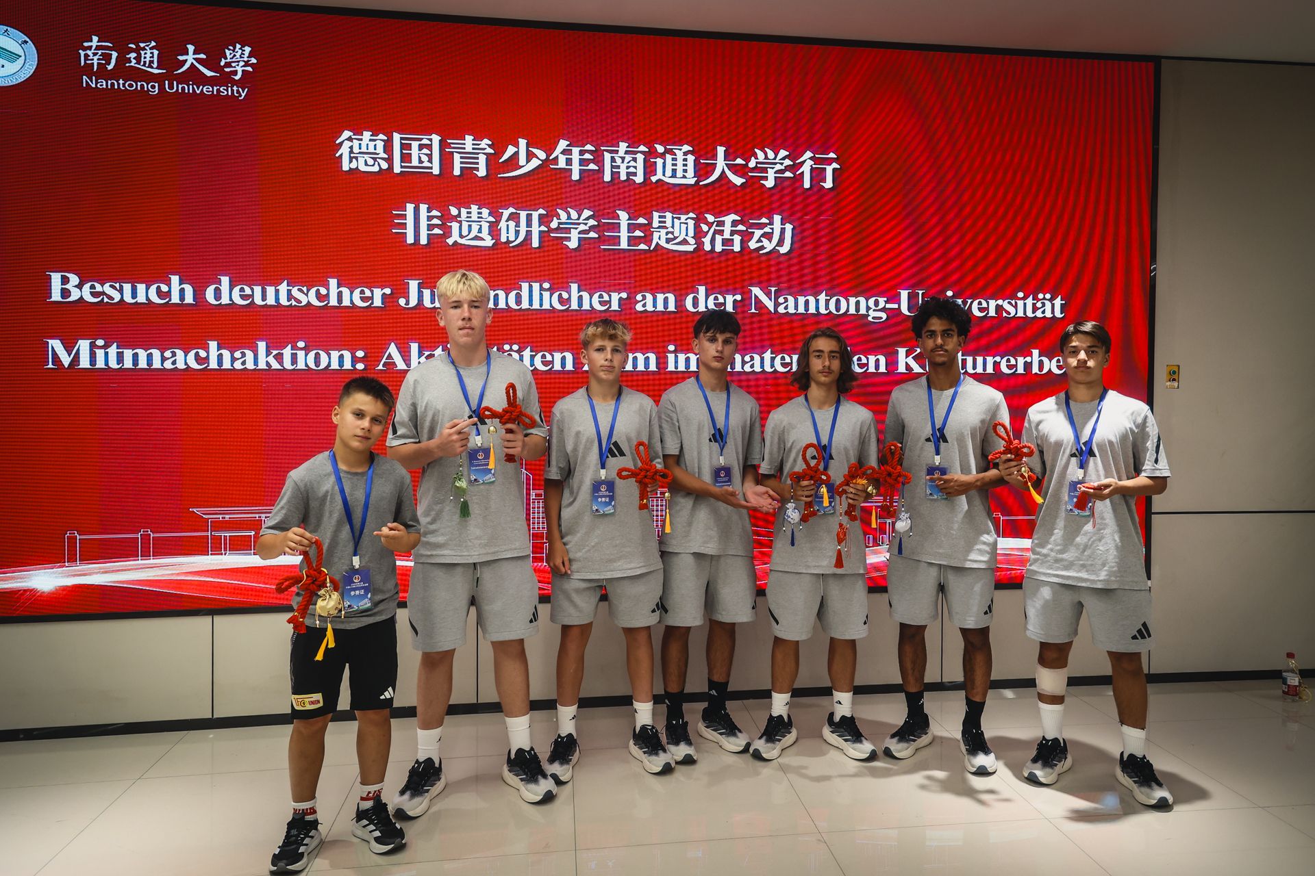 Gruppe junger Männer in Sportkleidung hält orangefarbene Schleifen und steht in einem modernen Raum mit Schildern der Nantong Universität.