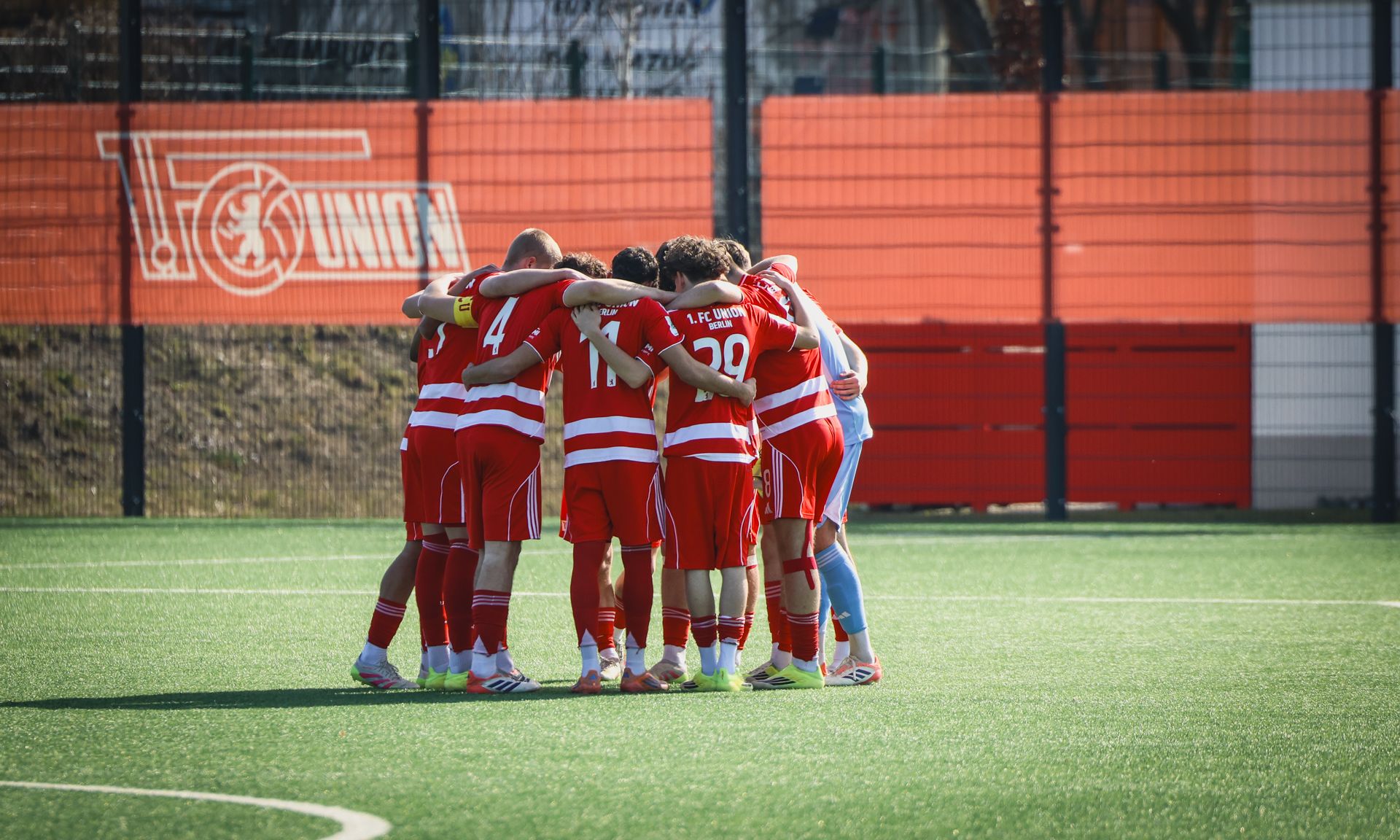 Eine Gruppe von Fußballspielern in roten Trikots steht eng zusammen und umarmt sich auf einem Sportplatz.