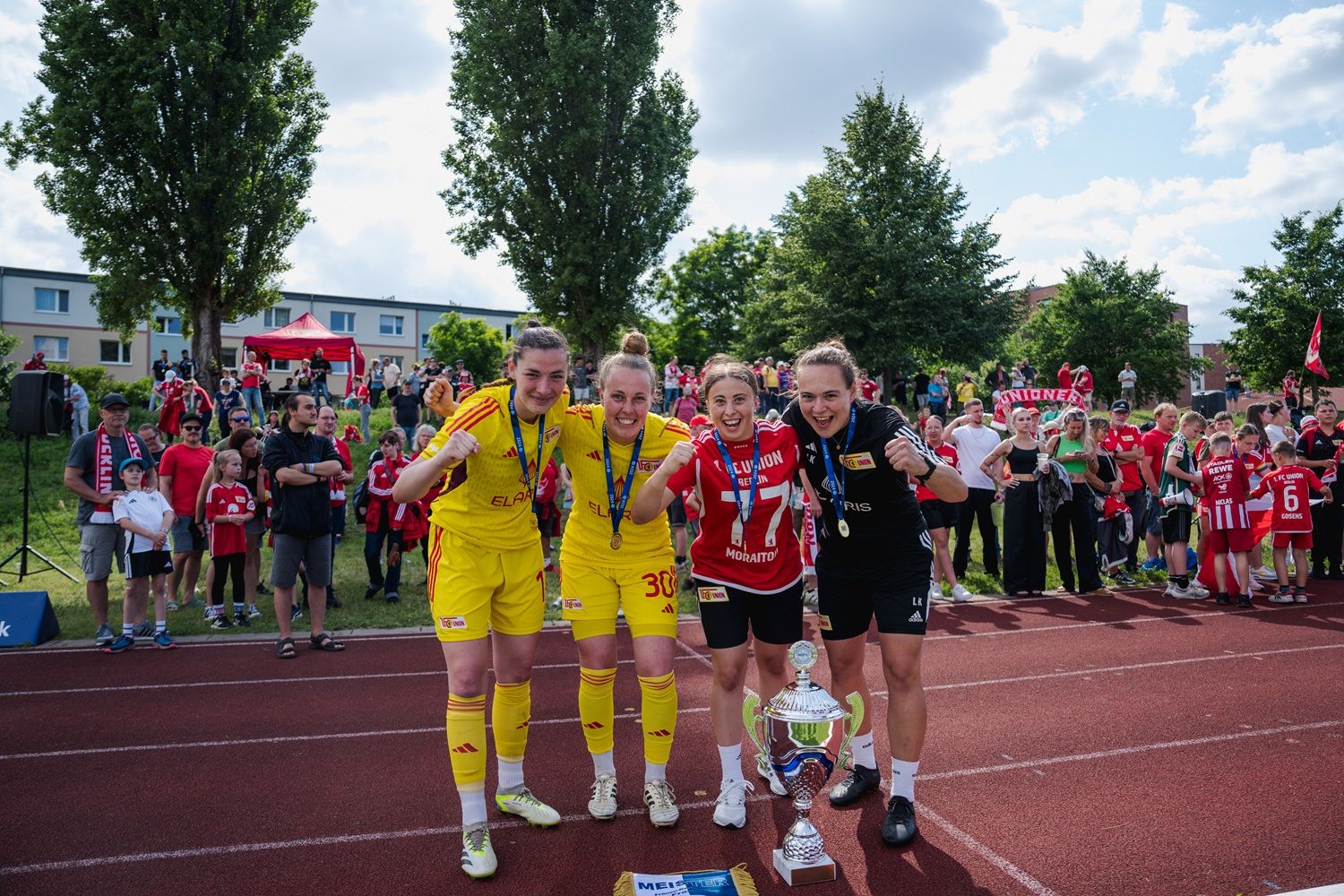 Vier Fußballspielerinnen posieren mit einem Pokal, umgeben von jubelnden Fans und einer festlichen Atmosphäre im Hintergrund.