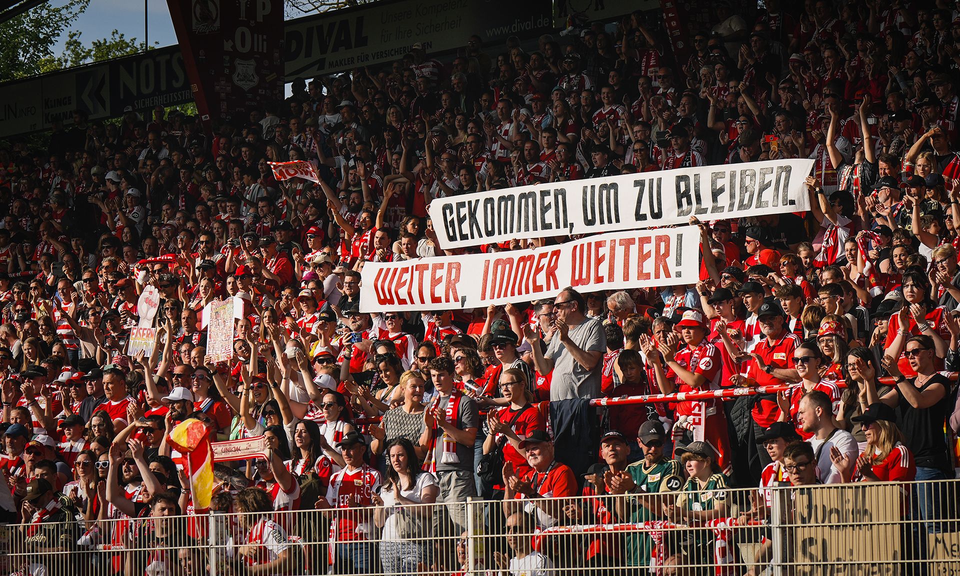 Zahlreiche Fans in einem Stadion stehen dicht gedrängt und halten Banner mit der Aufschrift „Gekommen, um zu bleiben. Weiter, immer weiter!“