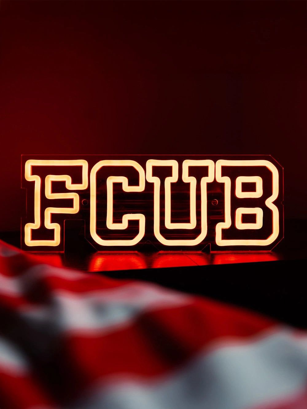 Leuchtendes Schild mit der Aufschrift "FCUB" vor einem roten Hintergrund und einem verschwommenen Flaggenmotiv im Vordergrund.
