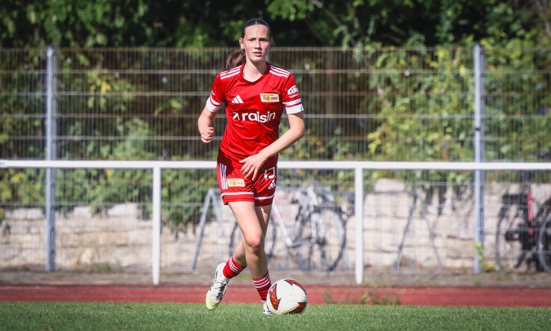 Eine Fußballspielerin in rotem Trikot und Shorts läuft auf dem Feld, umgeben von Bäumen und einer weißen Bande.