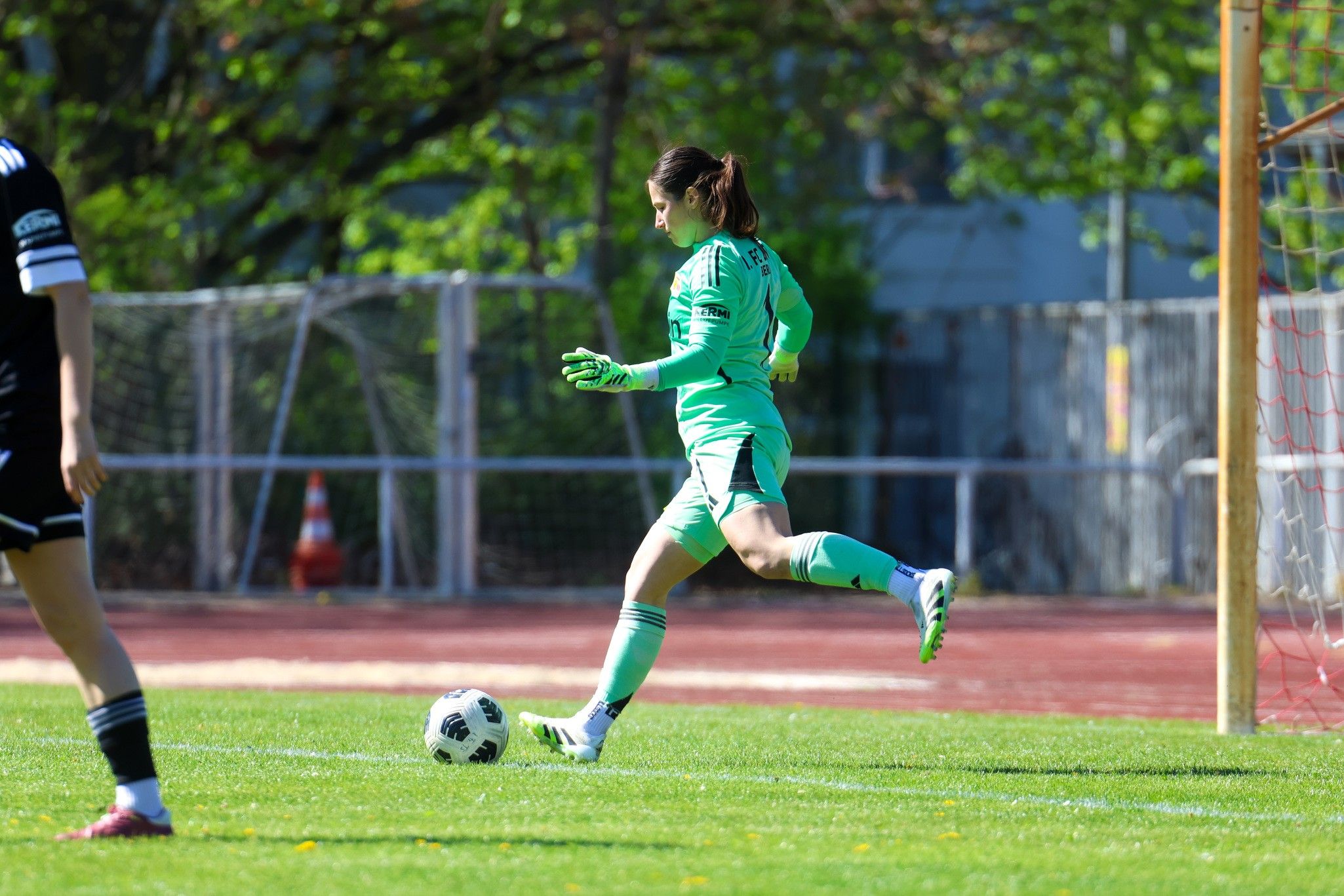 Torhüterin in grüner Ausrüstung tritt den Ball auf einem Fußballfeld. Hintergrund mit Bäumen und einem Tor.