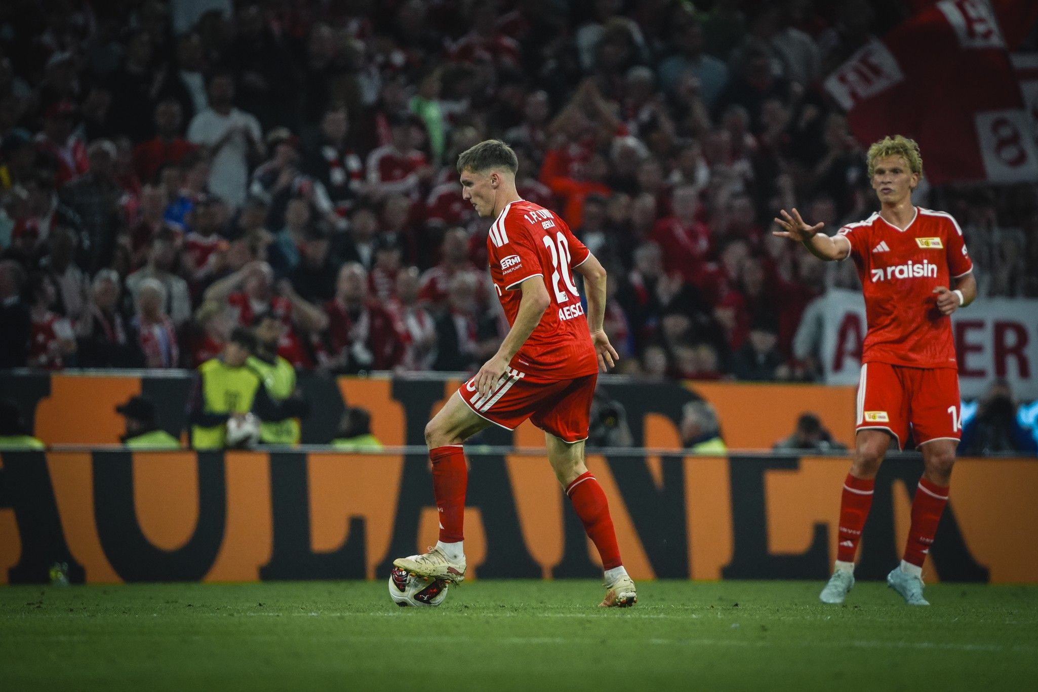 Ein Spieler in roter Fußballausrüstung steht auf dem Feld, während ein Teamkollege ihm gestikuliert. Im Hintergrund jubelnde Fans.