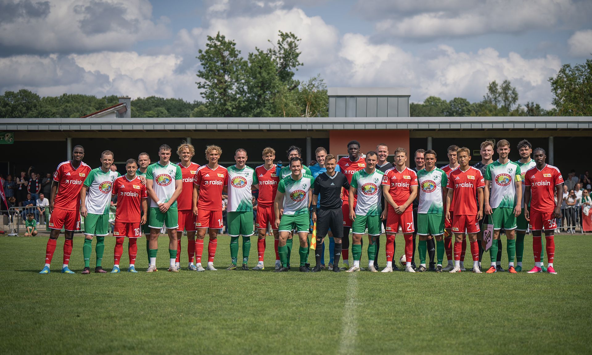 Zwei Fußballmannschaften stehen in Uniformen auf einem Rasenplatz, umgeben von Zuschauern und unter einem wolkigen Himmel.
