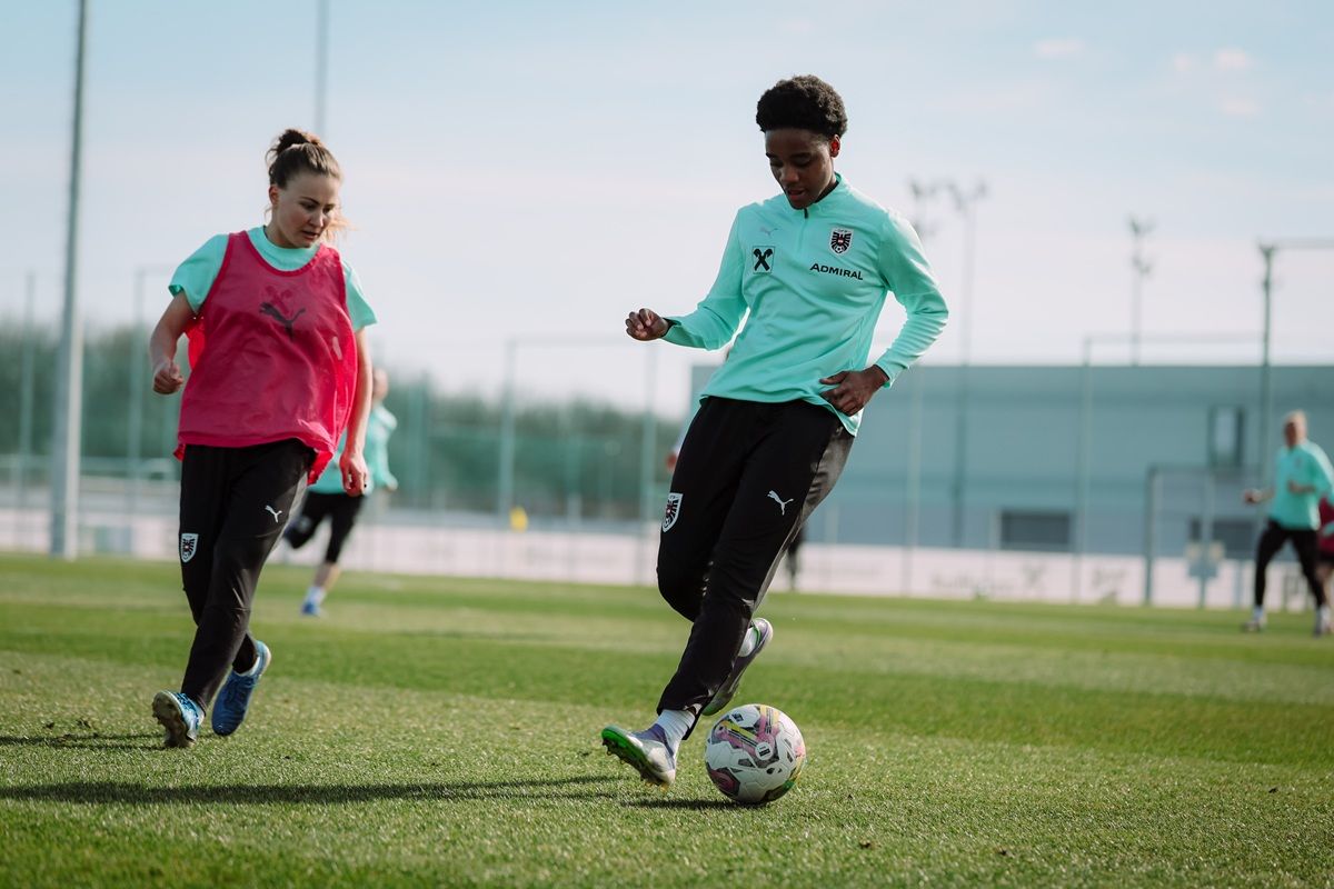 Zwei junge Fußballspielerinnen trainieren auf einem grünen Spielfeld, eine dribbelt den Ball, die andere verfolgt sie.
