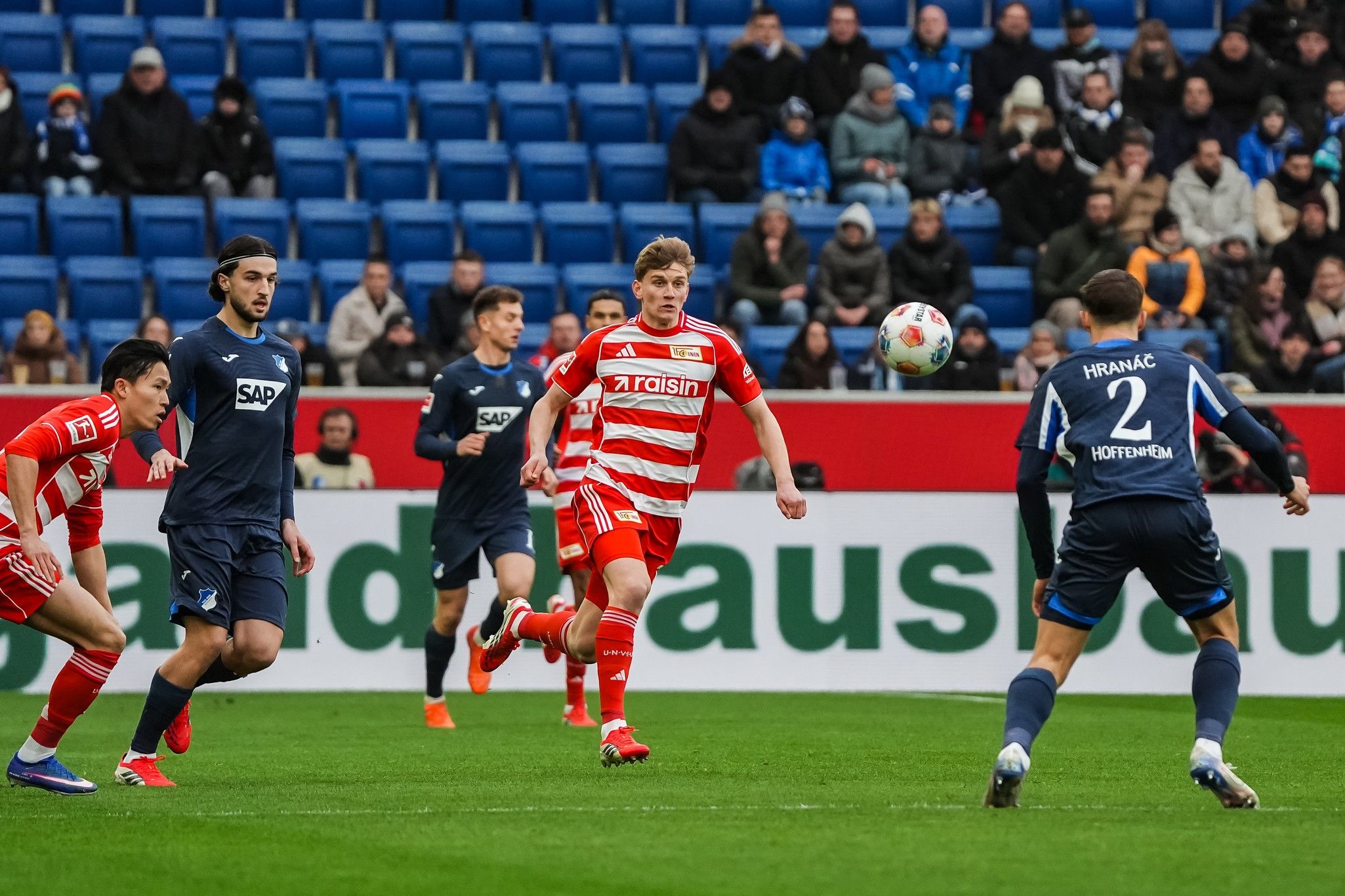 Ein Spieler in rot-gestreifter Uniform passt den Ball, während zwei Gegner in dunkler Kleidung in Angriffspostion stehen.