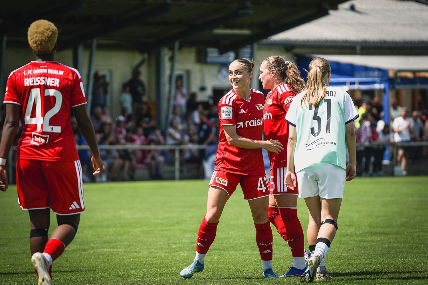 Vier Fußballspielerinnen auf dem Spielfeld, zwei in roten Trikots des 1. FC Union Berlin und zwei in weißen Trikots.