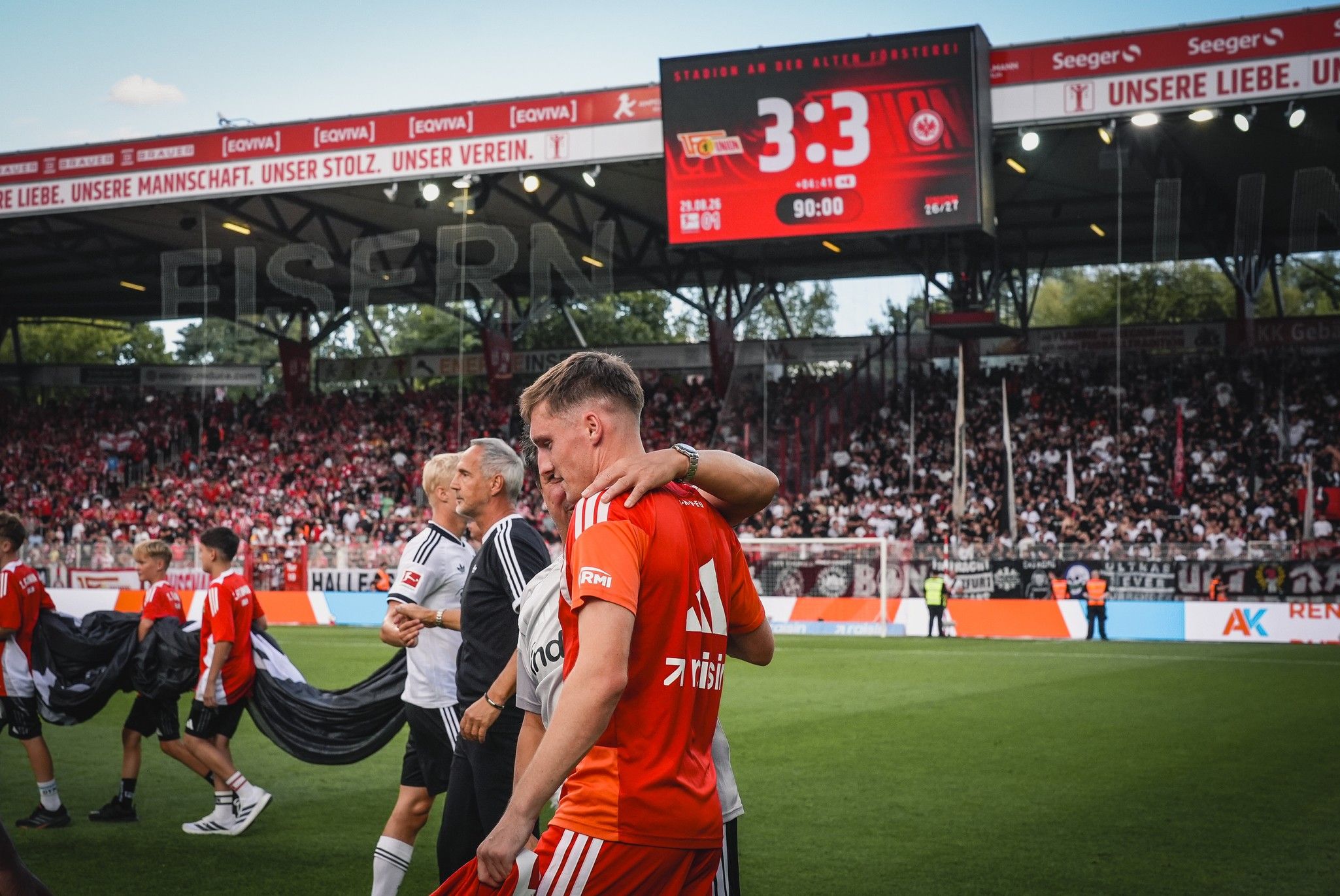 Zwei Fußballspieler feiern nach einem Spiel, im Hintergrund ein Stadion mit jubelnder Menge und einer Anzeigetafel mit 3:3.