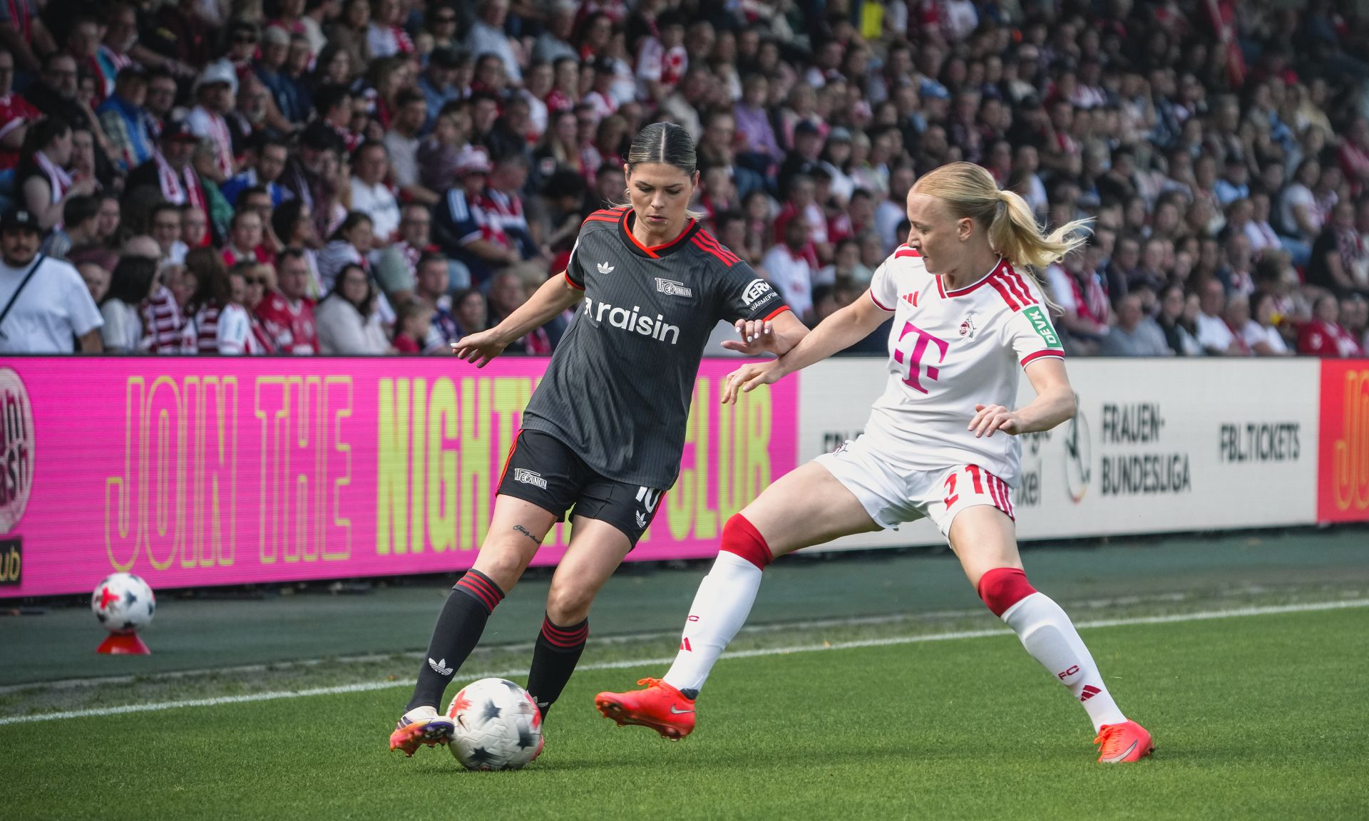 Zwei Fußballspielerinnen kämpfen um den Ball während eines Spiels, im Hintergrund jubelnde Zuschauer.