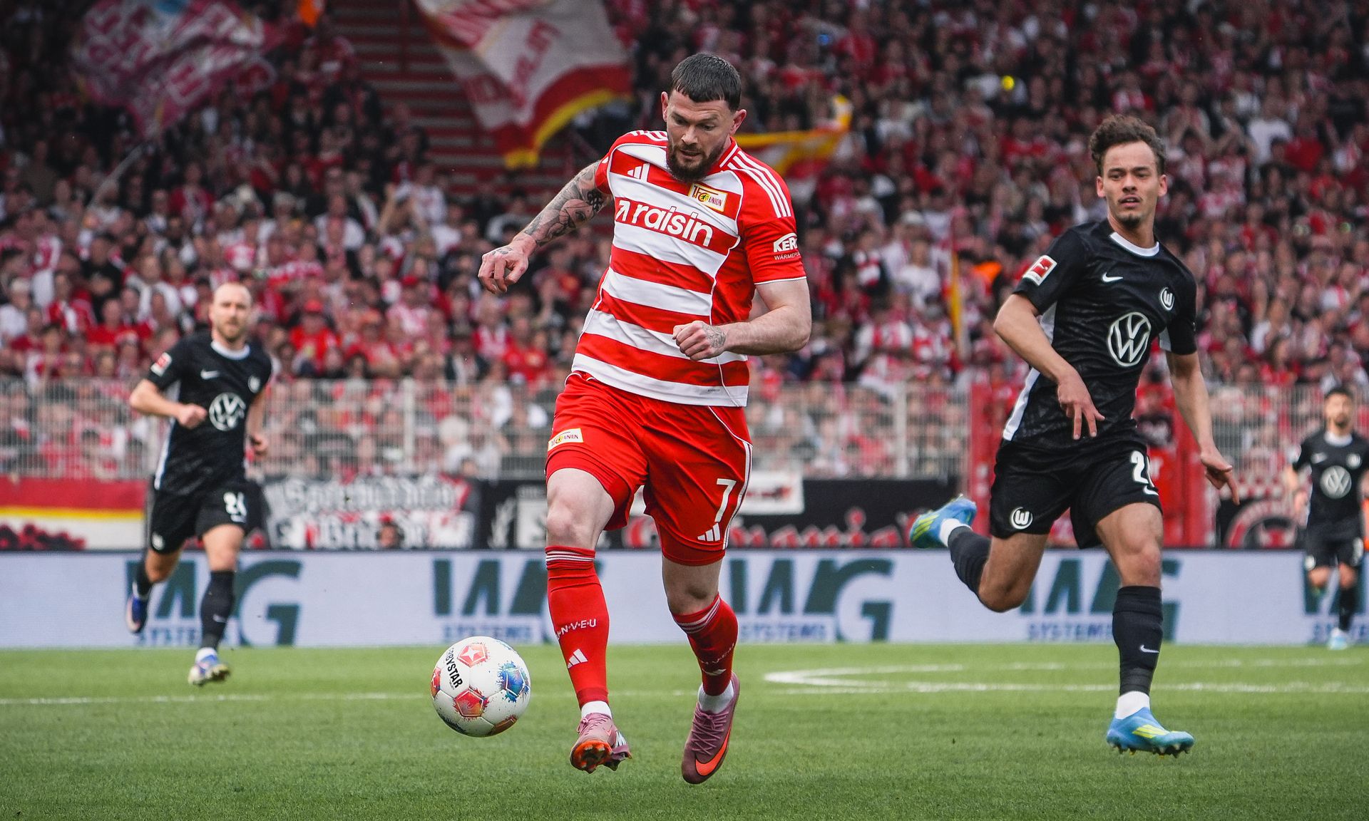 Ein Fußballspieler in gestreiftem Trikot läuft dem Ball entgegen, während ein Gegner ihn verfolgt. Im Hintergrund sieht man begeisterte Fans.