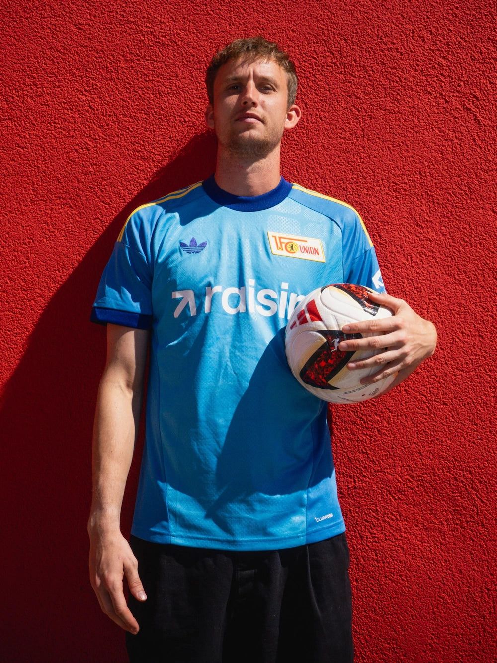 Ein junger Mann in einem blauen Fußballtrikot steht vor einer roten Wand und hält einen Fußball in der Hand.