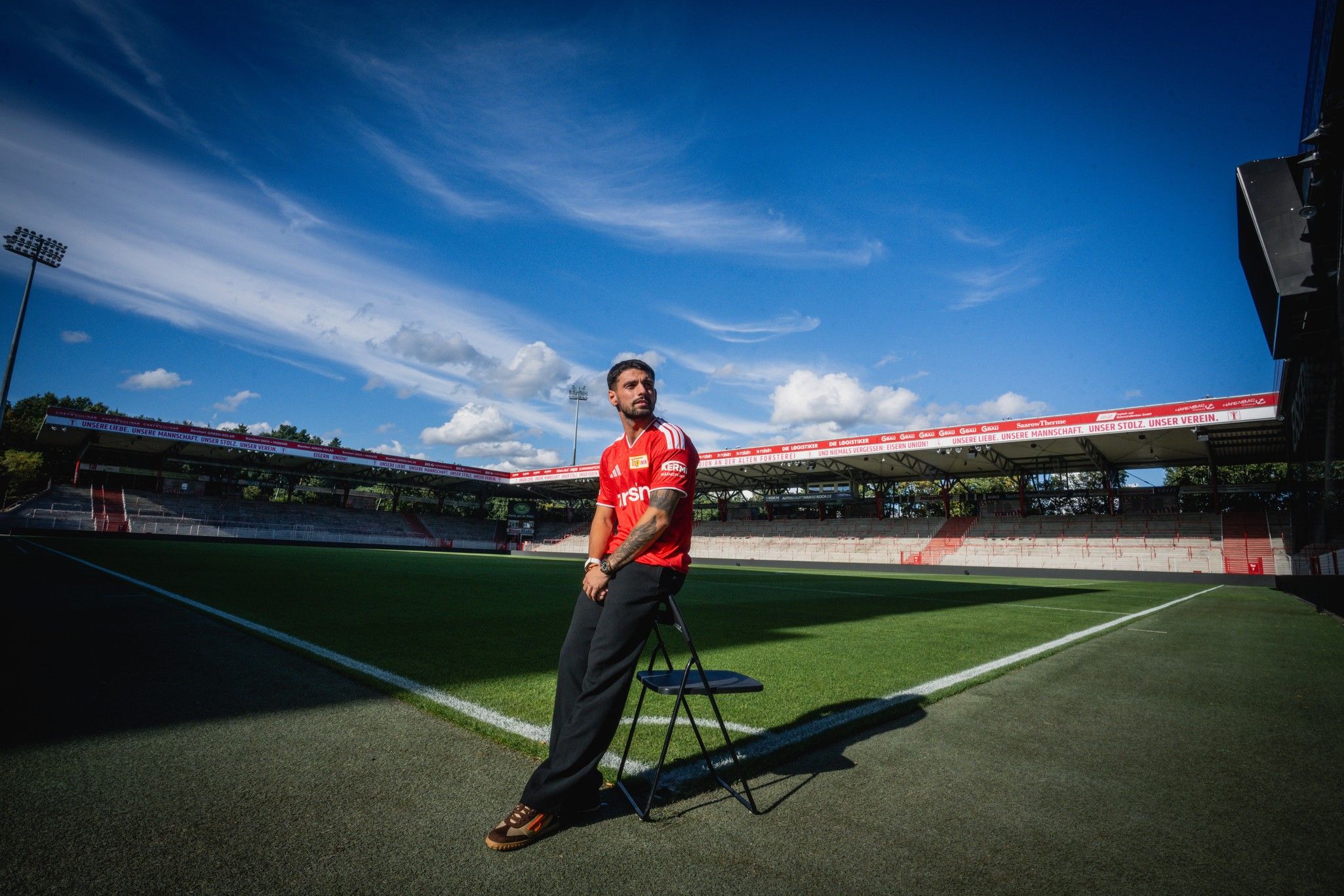 Ein Mann in rotem Trikot sitzt auf einem Stuhl im Stadion, umgeben von grünem Rasen und blauem Himmel.