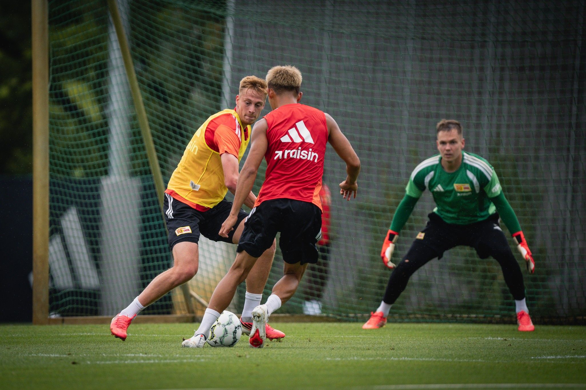Zwei Fußballspieler in roten Trikots trainieren auf dem Platz, während ein Torwart in grün hinter ihnen steht.