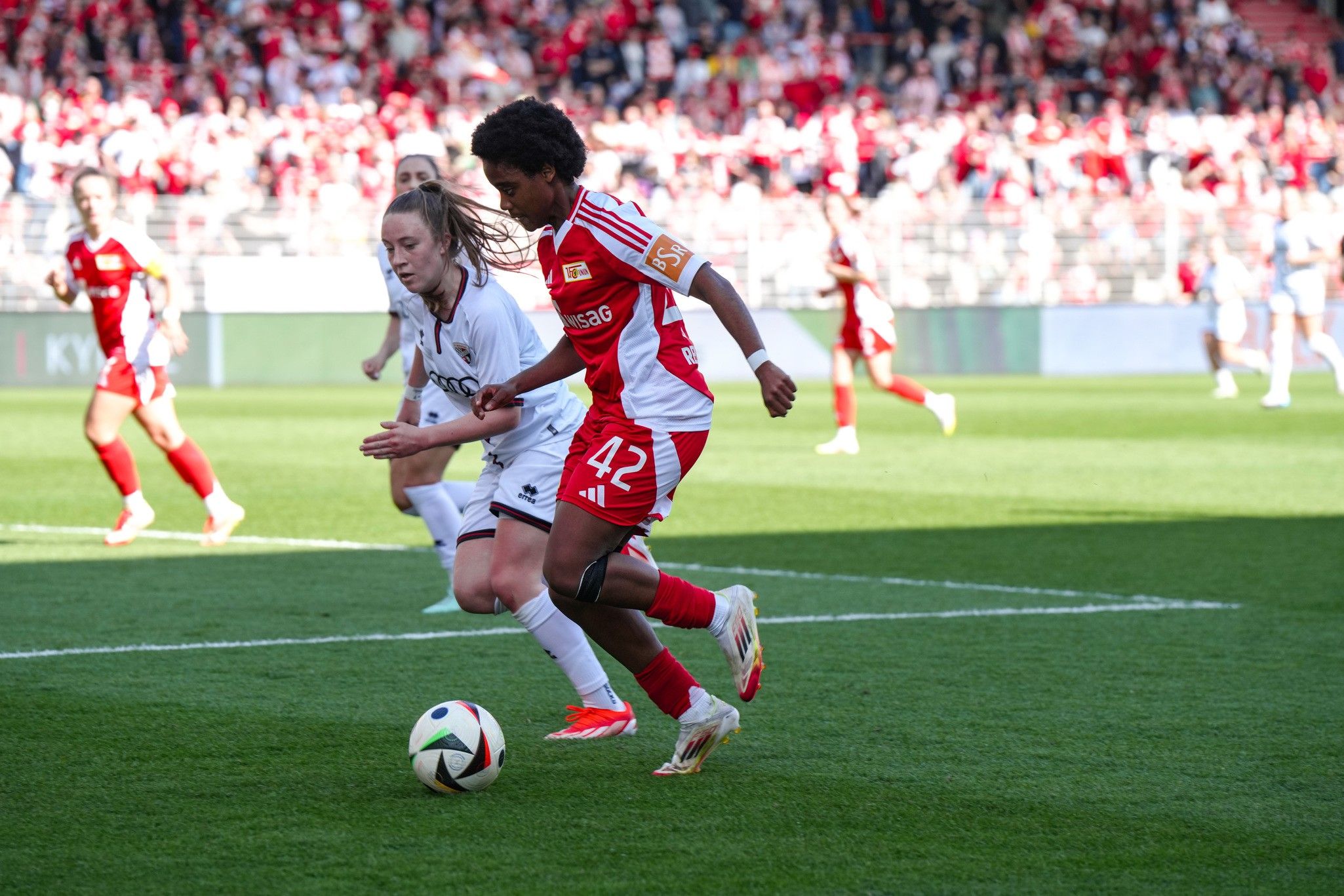 Zwei Fußballspielerinnen laufen um den Ball auf einem Spielfeld, mit Zuschauern im Hintergrund.