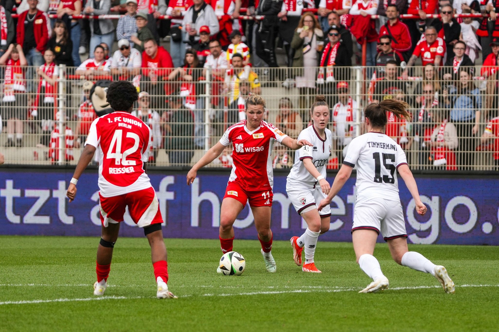 Szenen aus einem Fußballspiel: Spieler in roten Trikots stehen gegen Spieler in weißen Trikots, mit Zuschauern in einem Stadion im Hintergrund.