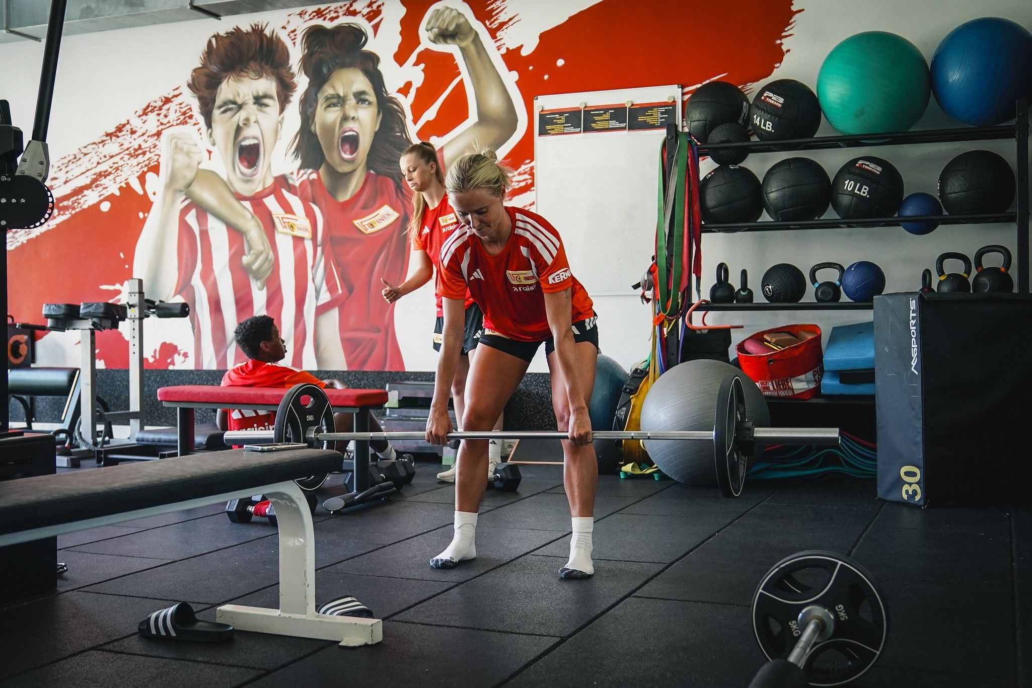 Eine Sportlerin hebt eine Hantel im Fitnessstudio, während andere Personen im Hintergrund trainieren und motivierte Wandbilder zu sehen sind.