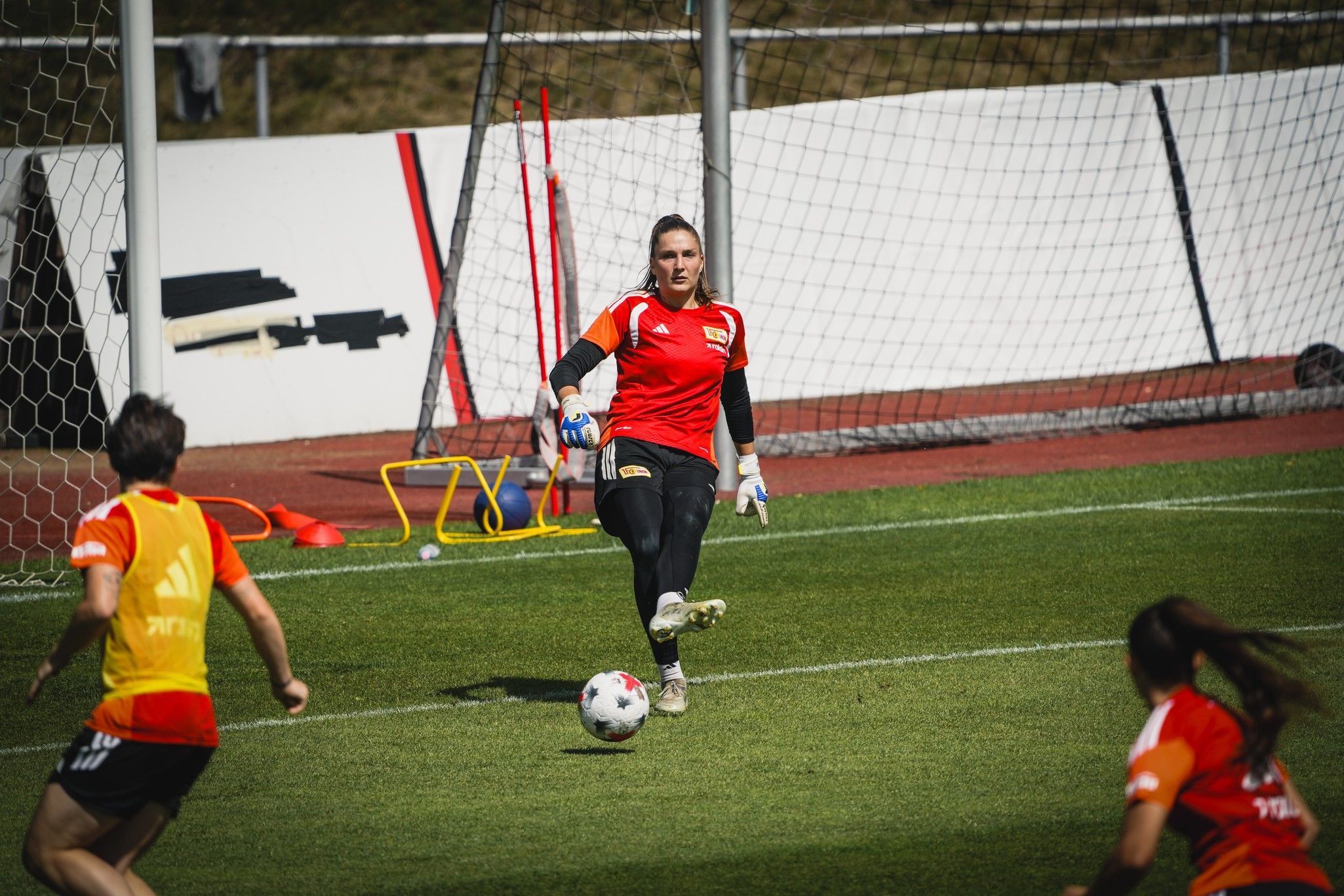 Torhüterin beim Training, bereit, den Ball zu spielen, während ihre Mitspieler sich im Hintergrund aufwärmen.