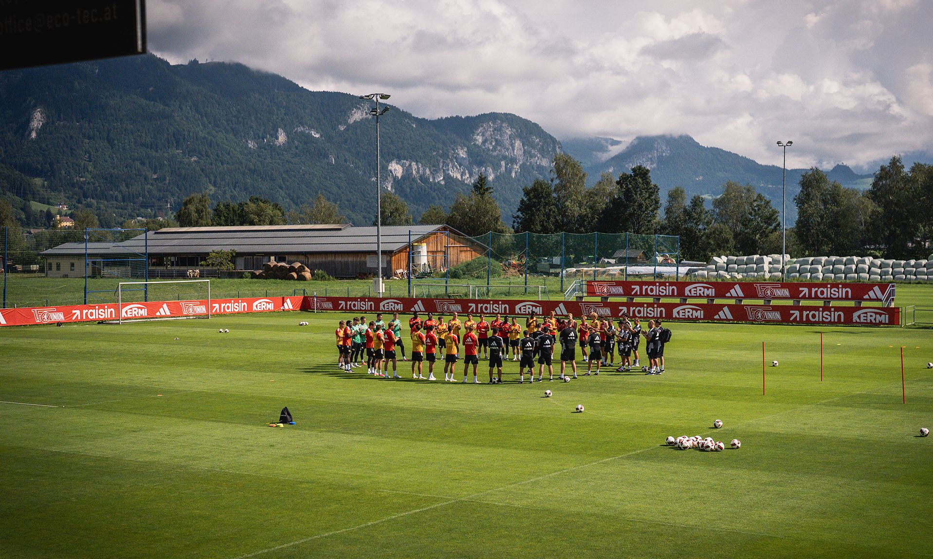 Gruppentraining einer Fußballmannschaft auf einem grünen Platz, umgeben von Bergen und leicht bewölktem Himmel.